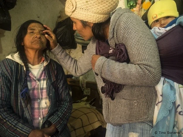 A woman helping an elderly woman with a basic eye examination in a rural setting.